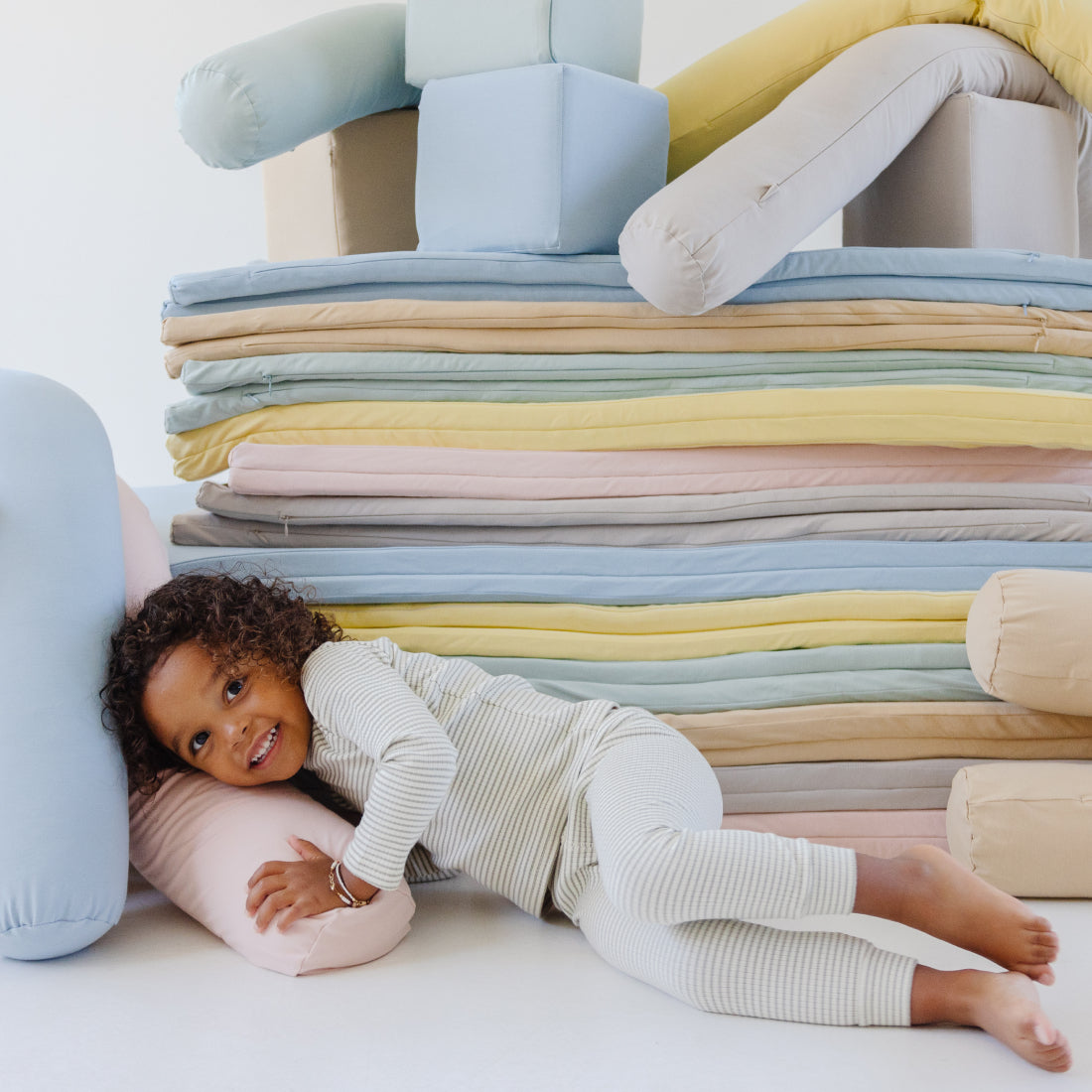 A child in striped pajamas joyfully leans on soft, multicolored cushions stacked in a cozy pile. The atmosphere is playful and comforting.