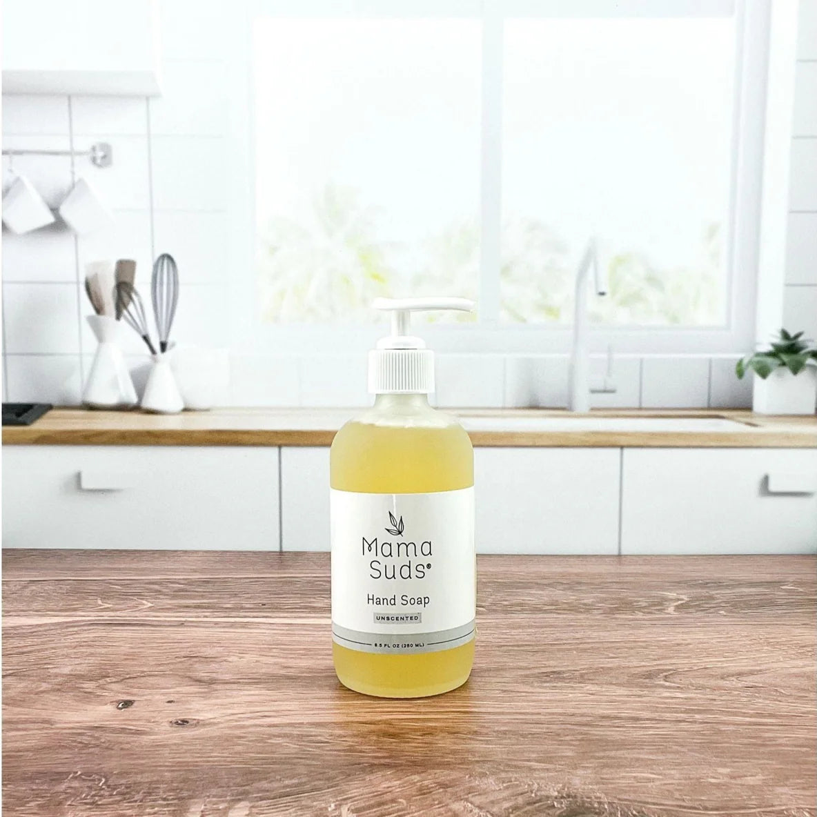 A glass bottle of liquid hand soap with a pump dispenser, labeled 'MamaSuds', placed on a wooden surface with a bright kitchen background.