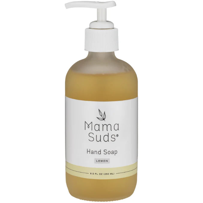 A glass bottle of liquid hand soap with a pump dispenser, labeled 'MamaSuds', placed on a wooden surface with a bright kitchen background.
