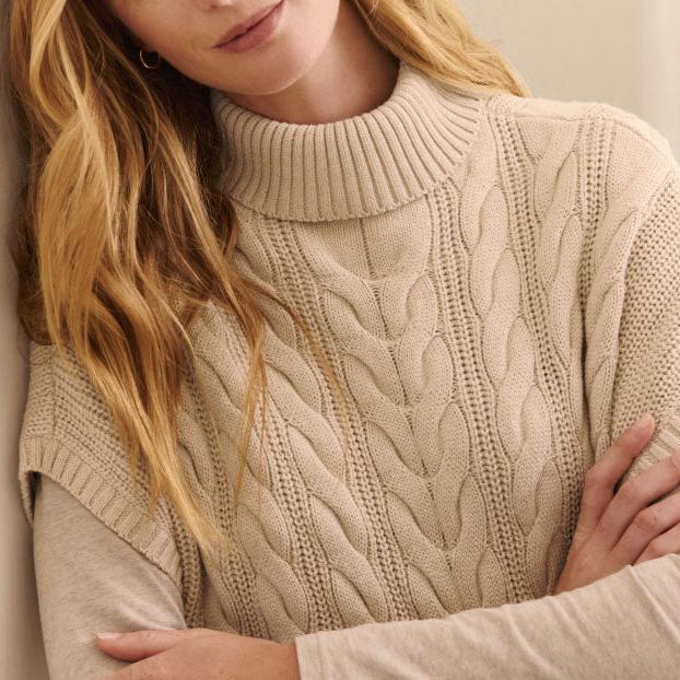 A woman with wavy blonde hair wears a beige, cable-knit turtleneck sweater layered over a light long-sleeve top, conveying warmth and coziness.