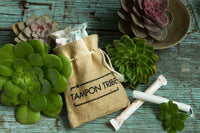 Burlap bag labeled 'Tampon Tribe' with tampons and succulent plants on a wooden surface