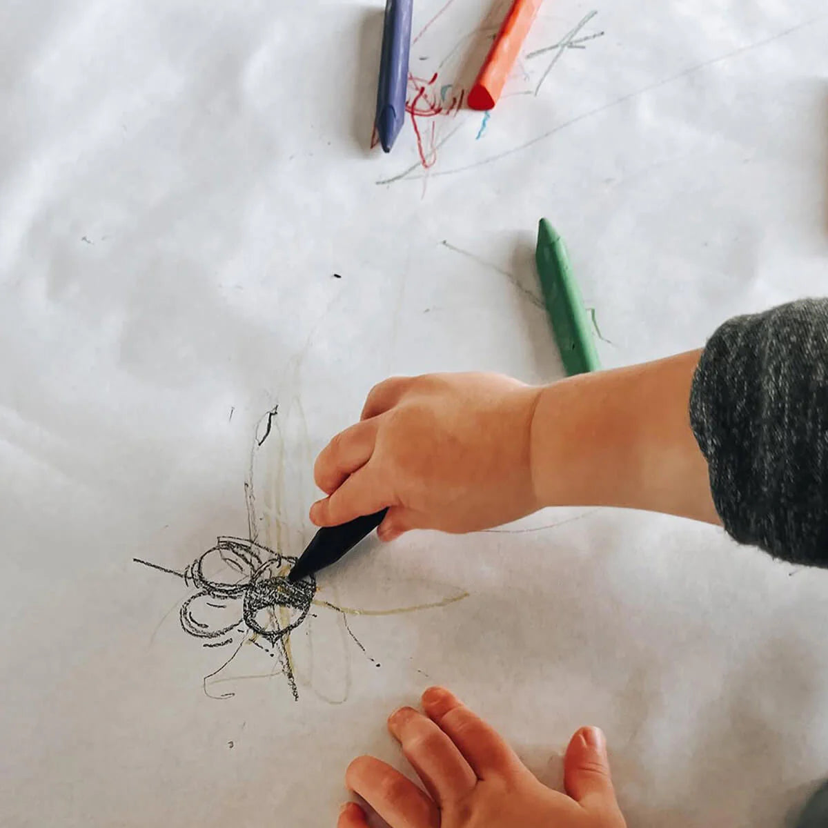 Child's hand drawing with a black marker on a white sheet of paper, with other markers visible.