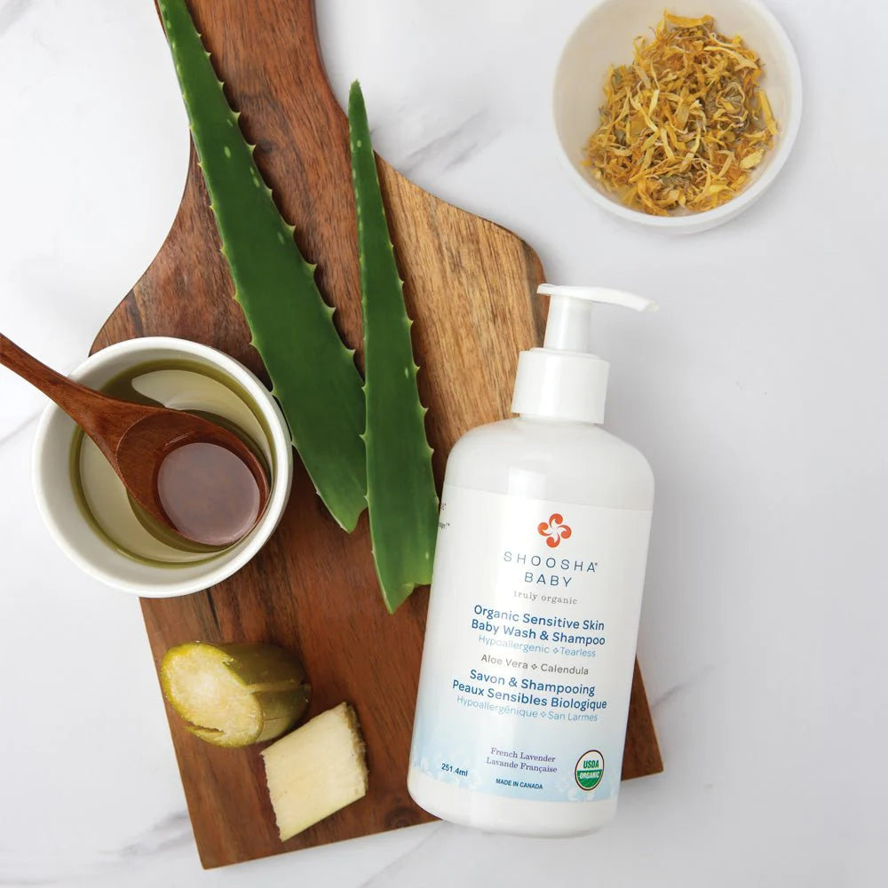 Bottle of organic baby wash on wooden board with aloe, ginger slices, bowl of oil with wooden spoon, and dried herbs, conveying a natural theme.