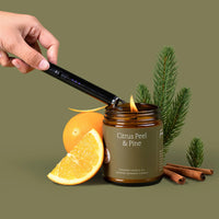 Hand lighting a candle labeled 'Citrus Peel & Pine' with oranges, cinnamon sticks, and pine branches on a green background.